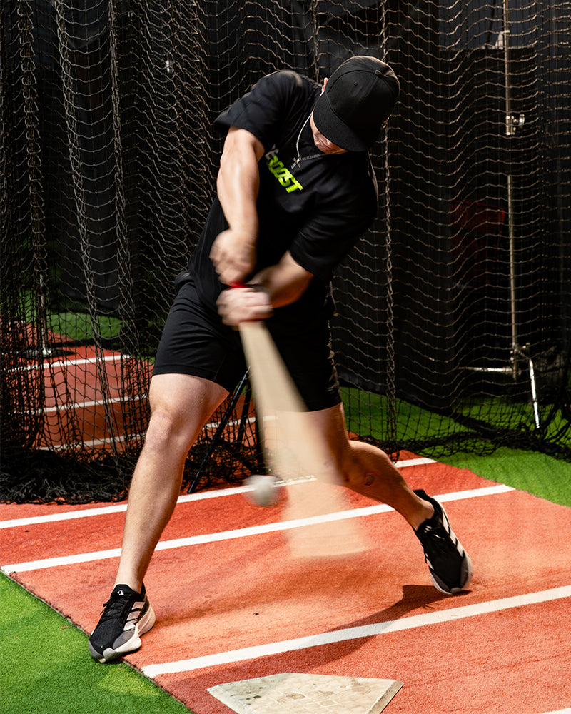 Jaxon Willits in a black t-shirt, black shorts, and a black cap swings a wooden baseball bat on an indoor hitting cage, standing on a red practice field with white lines.