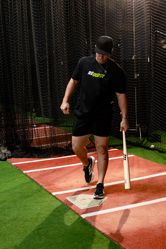 Jaxon Willits wearing a black t-shirt with a green and white REBOOST logo, black shorts, and a black cap walks home plate on an indoor baseball hitting range while holding a wooden bat.