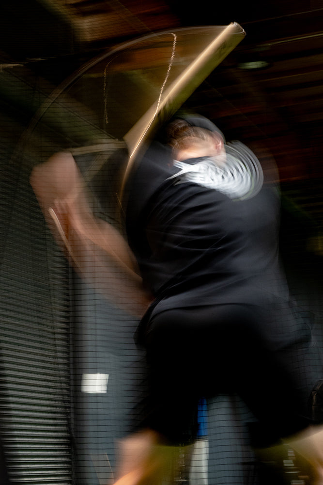 Jaxon Willits in a black outfit swinging a baseball bat indoors, captured with motion blur against a dark gray slatted wall.