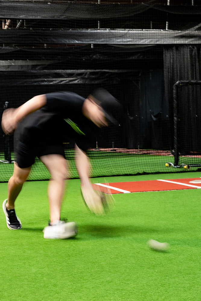 Jaxon Willits in a black t-shirt, black shorts, and a black cap is captured in motion on an indoor sports court with artificial turf, a baseball bat, and a ball, with a blurred effect to emphasize movement.