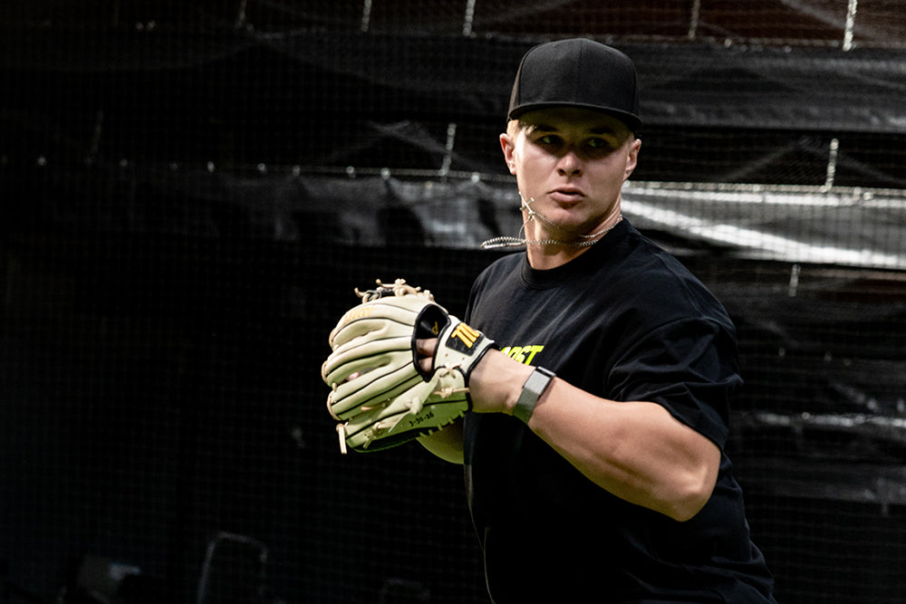 Baseball player Jaxon Willits in a black cap and black t-shirt holding a tan baseball glove, standing in front of a dark netting backdrop.