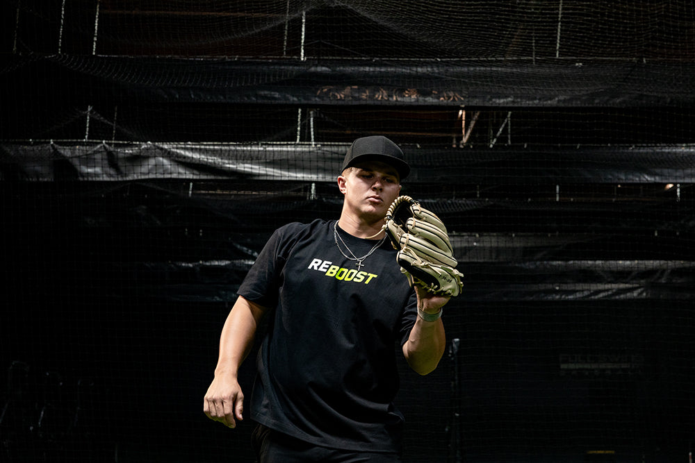 Jaxon Willits wearing a black t-shirt with 'REBOOST' printed in yellow and green on the chest, a black baseball cap, and a tan baseball glove, standing in front of a dark netted indoor sports facility backdrop.