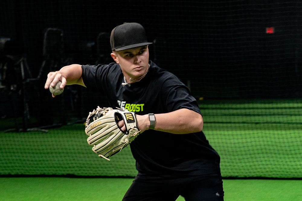 Jaxon Willits in a black t-shirt and cap throws a pitch on an indoor turf field, wearing a tan baseball glove and holding a baseball. The background shows a dark netting structure.
