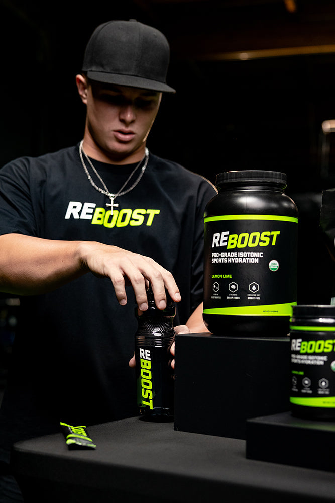 Jaxon Willits wearing a black REBOOST t-shirt and cap stands behind a counter, reaching for a REBOOST supplement bottle. Large black REBOOST tubs are displayed on black display blocks on the counter.