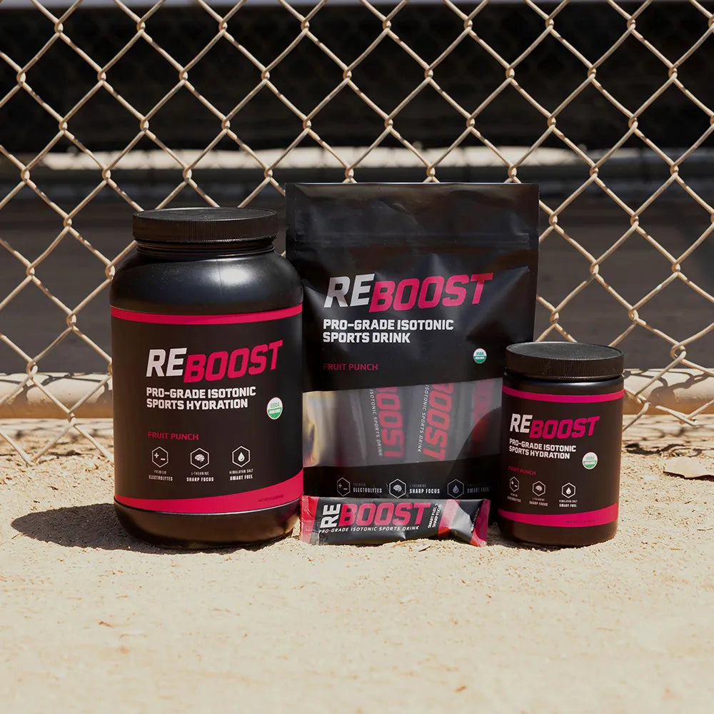 REBOOST fruit punch sports drink products on a sandy surface with a chain-link fence in the background