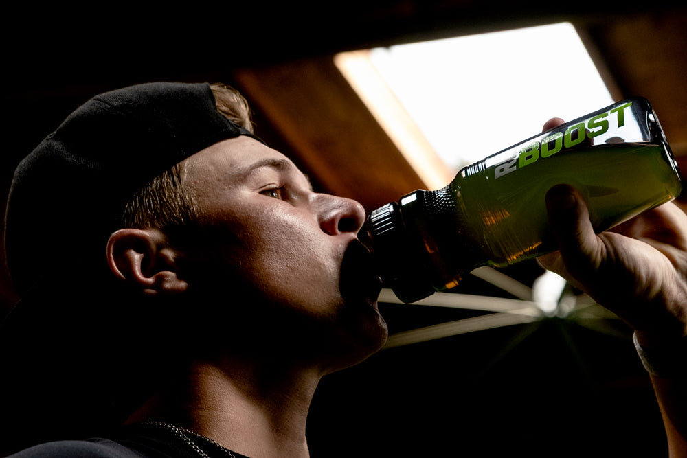 Jaxon Willits in a black cap drinking from a green and black bottle labeled 'REBOOST' in a dimly lit indoor setting.